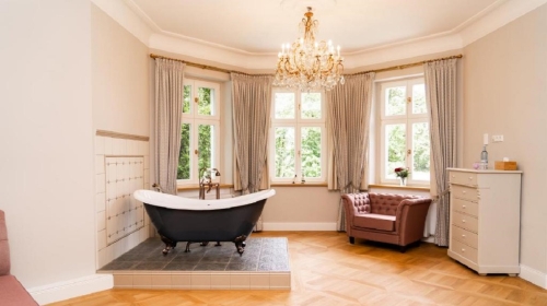 Diese Suite beeindruckt mit einer freistehenden Badewanne im Wohnbereich, gekrönt von einem stilvollen Kronleuchter. Chaiselongue, Sessel mit Fußhocker sowie ein großzügiger Eingangs- und Ankleidebereich schaffen ein luxuriöses Wohngefühl. 